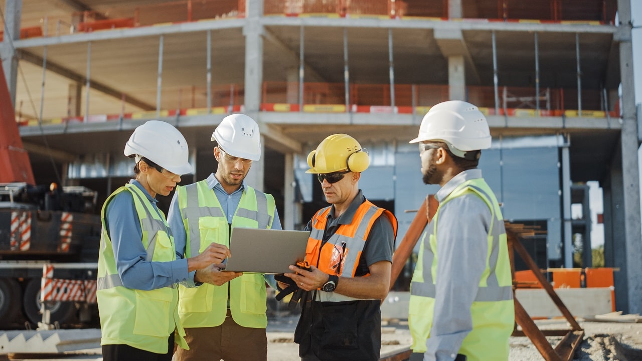 4 trabalhadores da construção civil com equipamentos de proteção seguram um tablet. Atrás deles, uma obra em andamento.