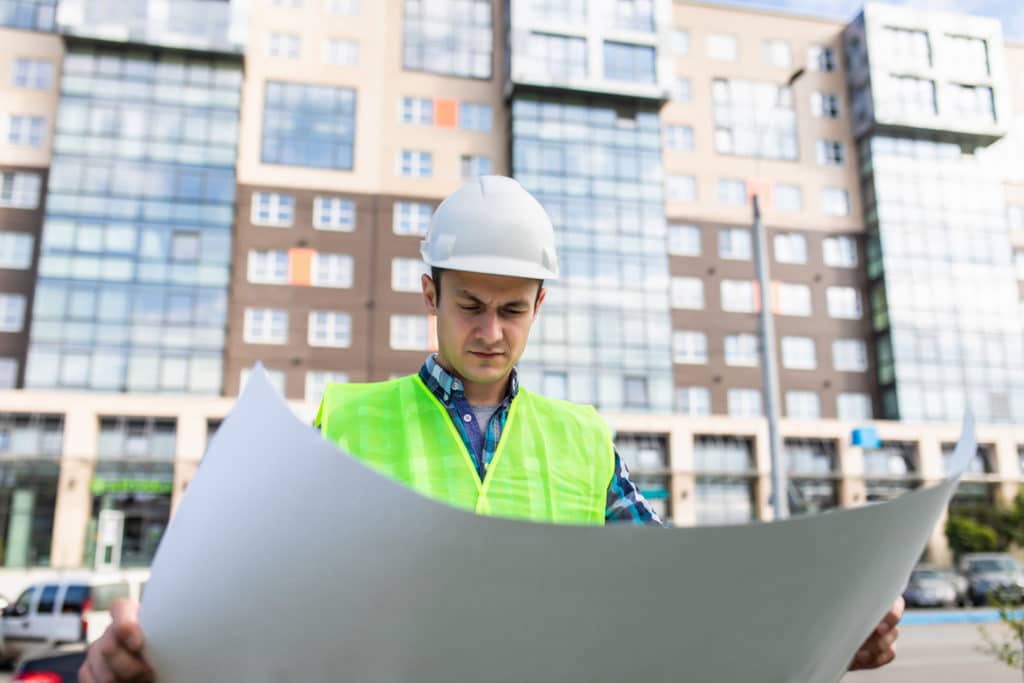 Homem olhando um projeto e atrás um conjunto de prédios.