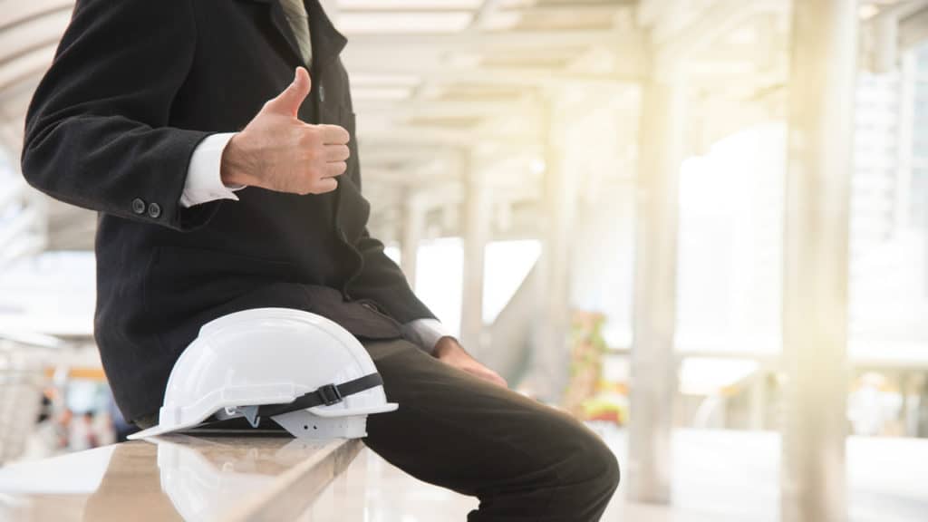 Homem de terno, fazendo o sinal de 'jóia' sentado ao lado de seu capacete de obras.