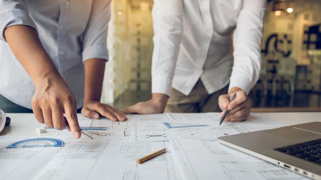 Dois arquitetos analisando plantas em cima de uma mesa e com um notebook ao lado